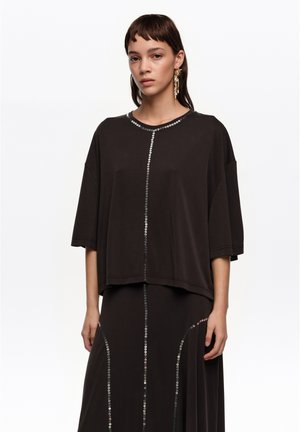 Joven mujer con blusa negra suelta y falda a juego con rayas verticales de lentejuelas, llevando pendientes largos y colgantes, de pie contra un fondo blanco.