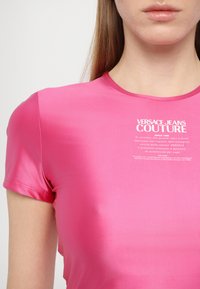 Fuksijas krāsas pieguļošs T-krekls, izgatavots no gludas, spīdīgas auduma ar īsām piedurknēm, ar baltu printētu zīmolu un kopšanas instrukcijām priekšpusē.