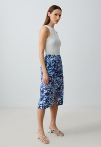 Top bianco senza maniche abbinato a una gonna a fiori blu fino al ginocchio con motivi geometrici e una fessura anteriore, indossata con sandali bianchi con tacco a blocco.