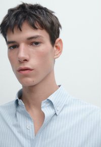 Camicia azzurra a righe con colletto, caratterizzata da una consistenza morbida e una sottile lucentezza del tessuto. Primo piano del viso di una persona.
