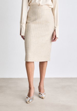 Beige woven pencil skirt with textured pattern, high waist, and knee-length. Paired with metallic two-tone heels featuring a pointed toe.
