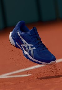 Chaussure de sport bleue avec des accents blancs, tige en maille respirante, semelle texturée et capuchon de orteil surélevé, capturée en plein vol au-dessus d'un court en terre battue.