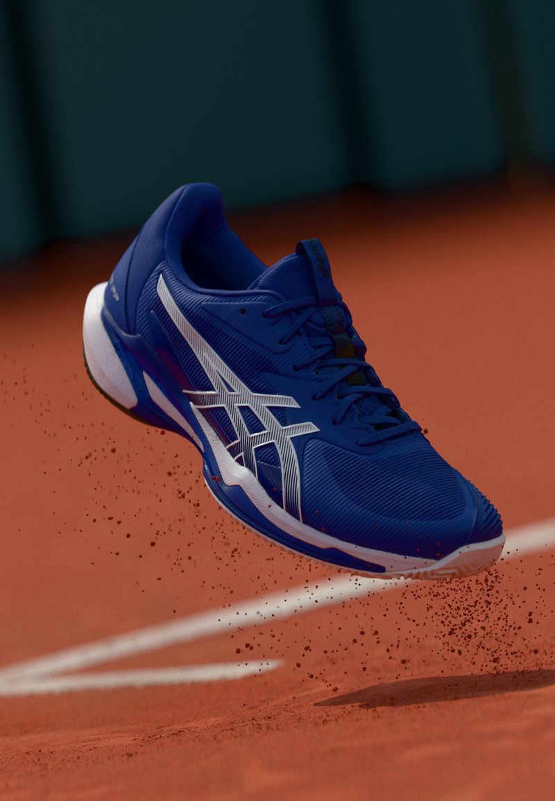 Chaussure de sport bleue avec des accents blancs, tige en maille respirante, semelle texturée et capuchon de orteil surélevé, capturée en plein vol au-dessus d'un court en terre battue.