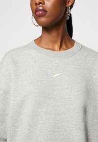 Gray sweatshirt made of soft fabric, featuring a crew neck and a small gold Nike logo embroidered on the chest. Textured detail in the neckline.