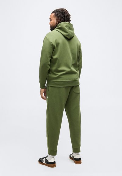 Άνδρας με πλεξουδάκια, ντυμένος με φούτερ και αθλητικά παντελόνια χλωρινού πράσινου χρώματος, στέκεται γυρισμένος προς τα πίσω σε λευκό φόντο.