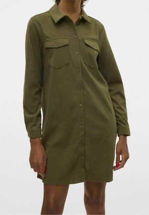 Donna che indossa un vestito camicia in velluto a coste color verde oliva con bottoni, due tasche sul petto e maniche lunghe.