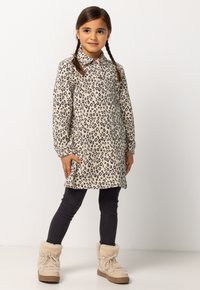 Jong meisje met gevlochten haar, gekleed in een beige jurk met luipaardprint, zwarte legging en beige winterlaarzen met bontvoering, staande tegen een witte achtergrond.