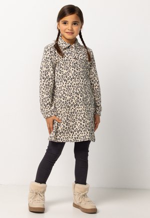 Jong meisje met gevlochten haar, gekleed in een beige jurk met luipaardprint, zwarte legging en beige winterlaarzen met bontvoering, staande tegen een witte achtergrond.