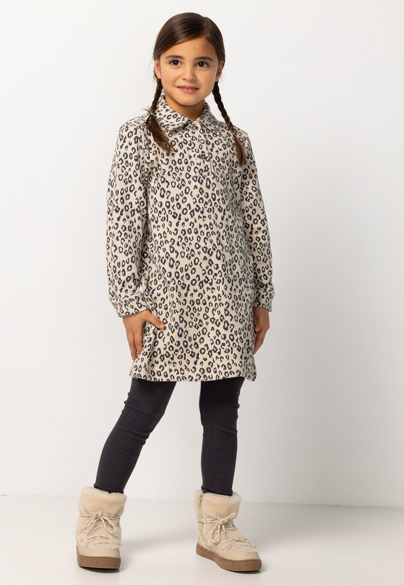 Jong meisje met gevlochten haar, gekleed in een beige jurk met luipaardprint, zwarte legging en beige winterlaarzen met bontvoering, staande tegen een witte achtergrond.