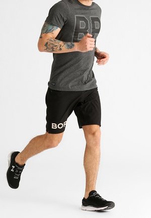 T-shirt de sport gris à manches courtes avec un logo foncé, associé à un short noir avec un texte "BORD" en blanc. Des chaussures de course noires complètent le look.