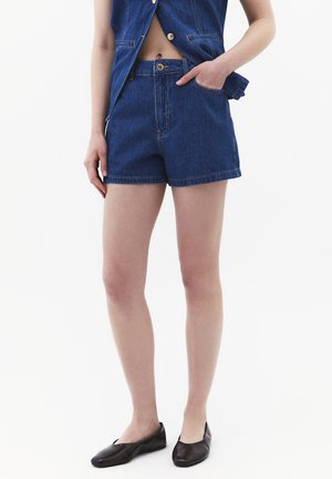 Person trägt hoch taillierte dunkelblaue Jeansshorts, passende geknöpfte Weste und schwarze flache Schuhe, Hand in der Tasche, steht vor weißem Hintergrund.