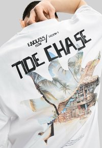 T-shirt bianca in cotone con una grande stampa grafica di una scena di spiaggia con palme e un capanno. Il testo "TIDE CHASE" appare sopra l'immagine.