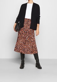 Svart stickad cardigan över en vit skjorta, kombinerad med en svart blommig midiklänning med orange och vita mönster, samt knähöga svarta stövlar.