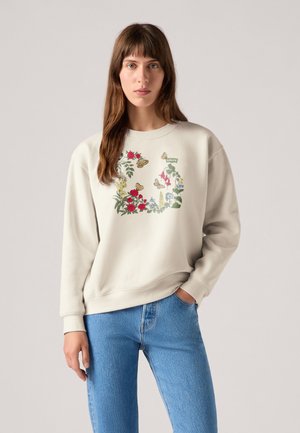 Ljusbeige sweatshirt med blommönsterbroderi i rött, grönt och blått. Har rundad halsringning och ribbade muddar, tillsammans med blå jeans.