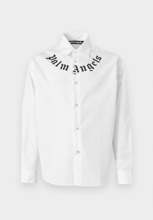 Homme portant une chemise blanche à manches longues avec le texte "Palm Angels" près du col, un short noir jusqu'aux genoux, des chaussettes noires et des baskets noires.