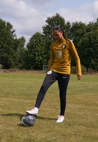 Soccer player in a gold long-sleeve top with logos, black pants, and white shoes, is poised to kick a patterned black and gray soccer ball on grass.