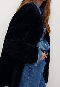 Zwarte fluffy jas met een zachte textuur, gedragen over een lichte spijkerhemd. Blauwe jeans en een zwarte riem maken het outfit compleet.