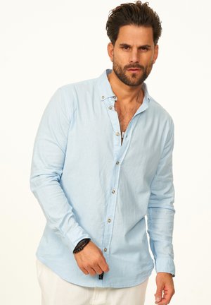 Bärtiger Mann mit gestyltem Haar, der ein hellblaues Hemd mit hochgekrempelten Ärmeln und weiße Hosen vor einfachem Hintergrund trägt.