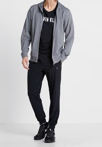 Sudadera con cremallera gris sobre una camiseta negra con el texto "CALVIN KLEIN", combinada con jogging negros y zapatillas deportivas negras. Tejidos suaves, corte casual.