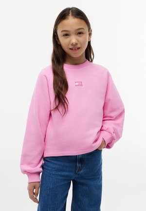 Κορίτσι με μακριά καστανά μαλλιά που φοράει ένα ροζ φούτερ και μπλε τζιν, στέκεται με το ένα χέρι στην τσέπη μπροστά σε ένα απλό φόντο.