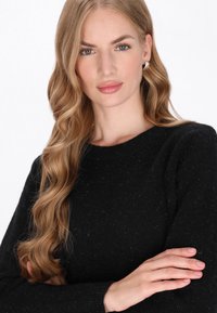 Suéter de punto negro con cuello redondo y mangas largas, con un tejido texturizado y sutiles motas. Acabado liso, ajuste holgado.