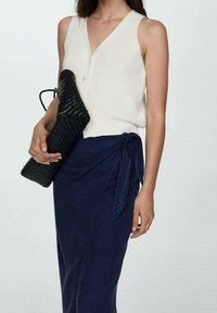 Crème gebreide tanktop met v-hals en knopen, gecombineerd met een marineblauwe wikkelrok met een gestrikte accent. Houdt een gestructureerde zwarte clutch vast.