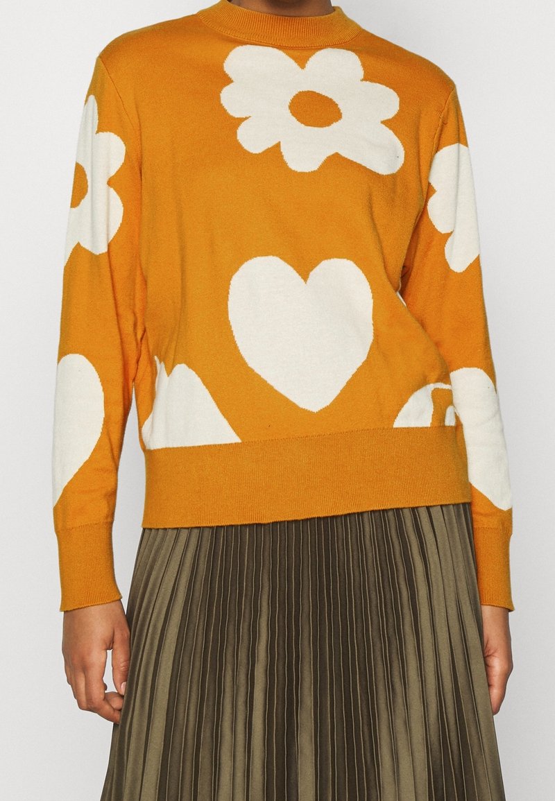 Pull orange avec des motifs de fleurs blanches et de cœurs, col rond, manches et ourlet côtelés. Associé à une jupe plissée vert olive.