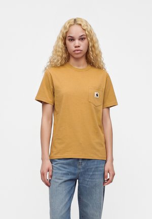 Jong persoon met lang, krullend blond haar draagt een mosterdgele T-shirt met zakje en blauwe spijkerbroek, staand tegen een effen achtergrond.