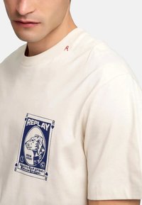 Camiseta color crema con un estampado gráfico azul que presenta un león y la palabra "REPLAY". Detalle bordado rojo en el cuello. Material de algodón.