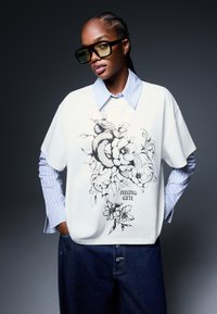 Bela oversize majica z black floral serpentinim grafičnim dizajnom in napisom "FEELING CUTE", prek večslojne modro črtaste srajce, v kombinaciji z temnimi kavbojkami.