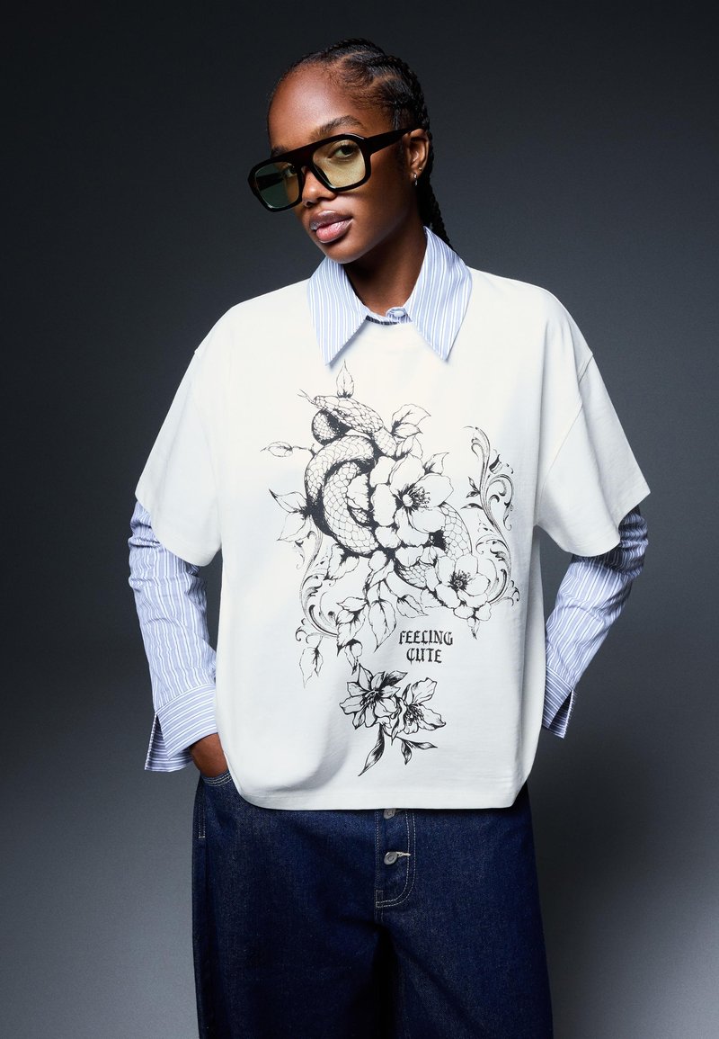 Bela oversize majica z black floral serpentinim grafičnim dizajnom in napisom "FEELING CUTE", prek večslojne modro črtaste srajce, v kombinaciji z temnimi kavbojkami.