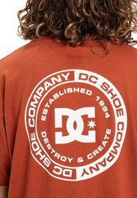 Tričko z bavlny v rustikálnej farbe s veľkým bielym kruhovým logom v strede, na ktorom je nápis "DC Shoe Company" a "Destroy & Create".