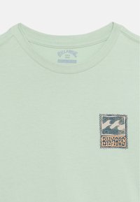 Világoszöld, kerek nyakú póló, amelyen apró, négyzet alakú Billabong logó látható a mellrészen, két stilizált óceáni hullámmal és a márkanévvel.