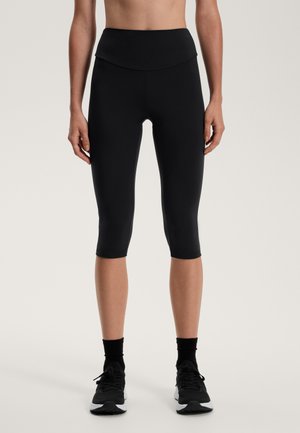 Fekete capri leggings, magas, hajtott derékkal. Rugalmas anyagból készült, sima textúrájú, és fekete sportcipővel vannak párosítva.