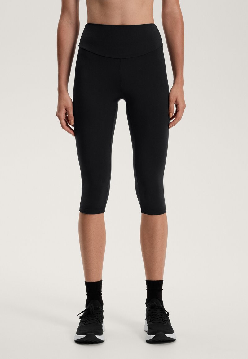 Czarne legginsy capri z wysokim, złożonym pasem. Wykonane z elastycznego materiału, mają gładką fakturę i są zestawione z czarnymi butami sportowymi.