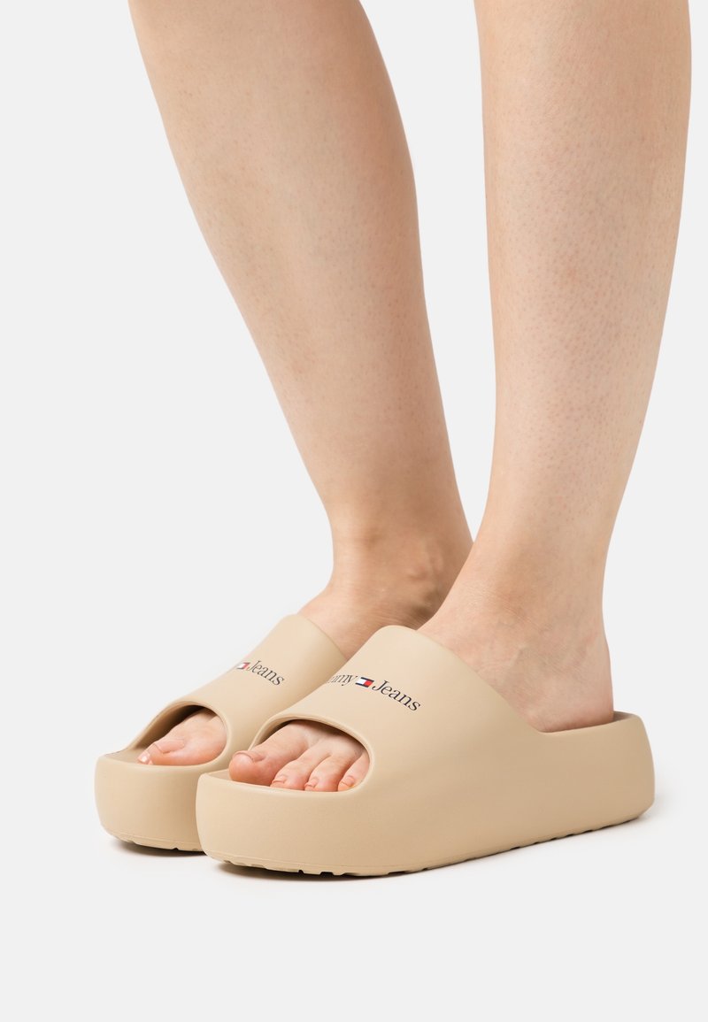 Pieds portant des sandales à plateforme beige avec le logo Tommy Jeans sur les brides, montrés sur un fond blanc uni.