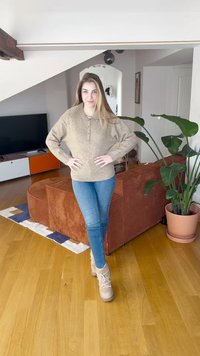 Šviesiai rudas megztinis su sagotu iškirptės kaklu, mėlyni aptempti džinsai ir smėlio spalvos sportbačiai. Fone matyti ruda sofa ir vazoninis augalas.