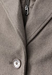 Grauer Wollmantel mit Schalkragen, Reißverschluss und einer metallischen Knopf mit graviertem Text. Weicher Griff und taillierte Passform.