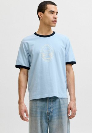 Junger Mann trägt ein hellblaues T-Shirt mit dunklem Saum und weißem grafischem Text, kombiniert mit locker sitzenden hellen Jeans vor schlichtem Hintergrund.