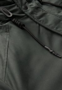 Mörkgrön vattentät jacka med en slät yta, prydd med svarta dragsnören, knappar och en elegant design. Nära bild av detaljer.