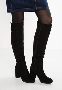Botas negras hasta la rodilla hechas de suave ante, con tacón cuadrado, cierre de cremallera y una textura lisa. Acentuadas con costuras mínimas.