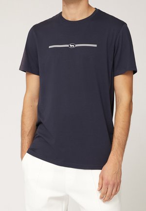 Man wearing navy blue short-sleeve T-shirt with white horizontal stripe and small dog logo, paired with white pants.