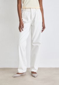 Pantalones de denim blanco con corte relajado, que presentan una cintura alta, bolsillos frontales y costuras visibles. Combinados con zapatos planos de color gris claro.