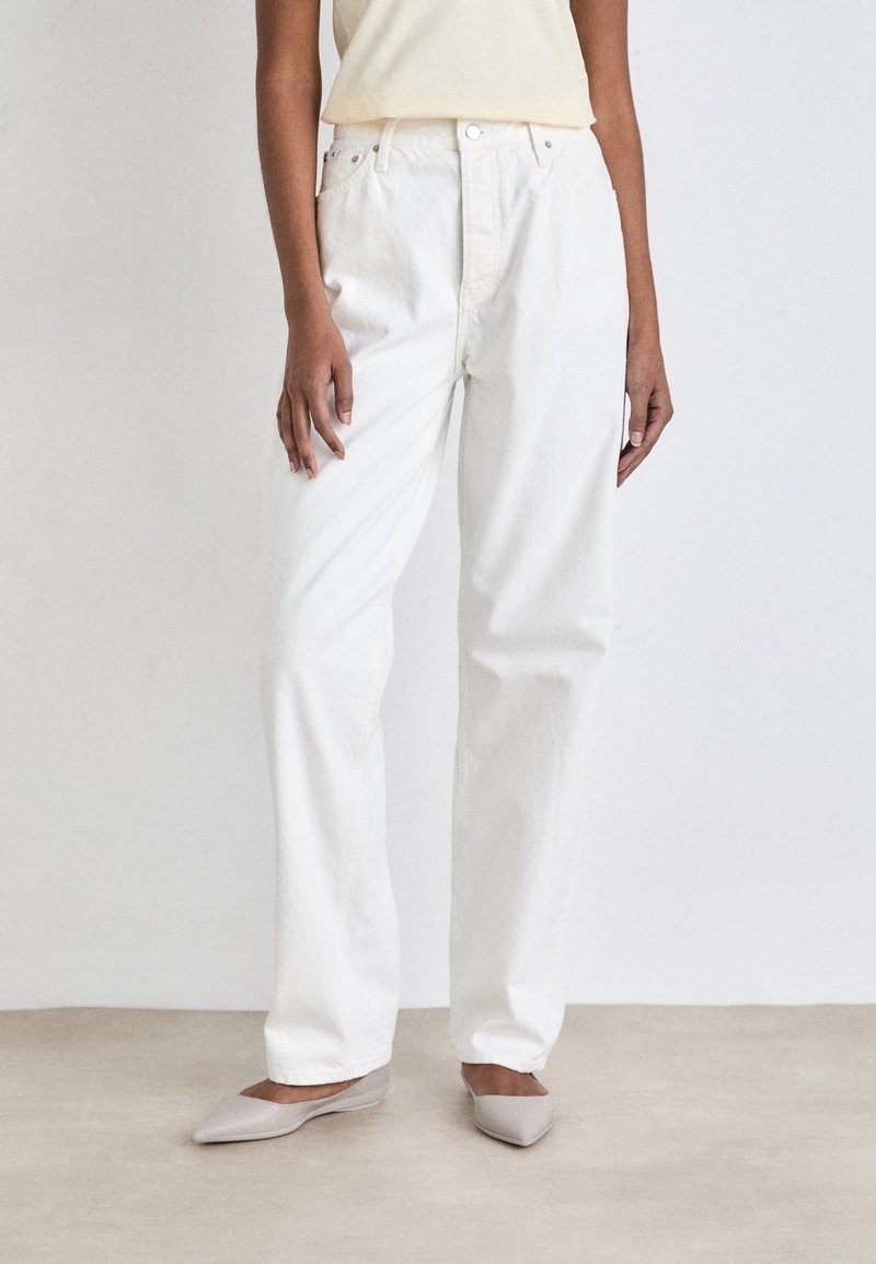 Pantalones de denim blanco con corte relajado, que presentan una cintura alta, bolsillos frontales y costuras visibles. Combinados con zapatos planos de color gris claro.