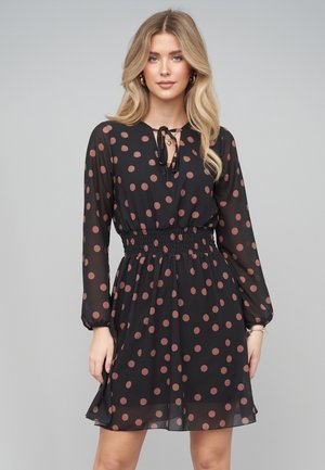 Frau mit langen blonden Haaren trägt ein schwarzes, braunes gepunktetes, langärmliges Kleid mit geraffter Taille und Schleifenbindehals auf grauem Hintergrund.