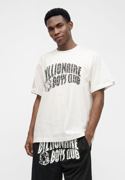 Νέος άνδρας που φοράει λευκό T-shirt Billionaire Boys Club και μαύρα σορτς με το ίδιο λογότυπο, στέκεται με το ένα χέρι στην τσέπη.