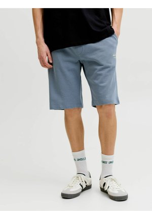 Mężczyzna w niebieskich, casualowych szortach, białych skarpetkach z zielonym napisem, białych i szarych trampkach oraz czarnej koszulce z jedną ręką w kieszeni.