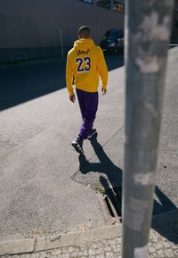 Moletom amarelo com "James 23" nas costas, combinado com calças roxas e sapatos pretos. Cenário urbano com um poste de metal parcialmente visível.