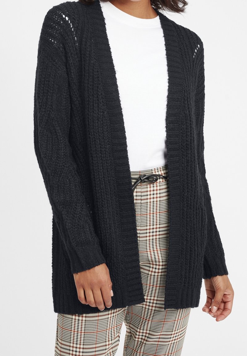 Svart stickad cardigan med öppen framsida, ribbat mönster och långa ärmar, matchad med rutiga högmidjade byxor.