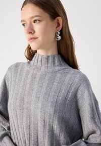Šedý žebrovaný svetr s vysokým límcem, se svislými pruhy. Stříbrné kulaté náušnice doplňují design. Měkká textura je patrná.